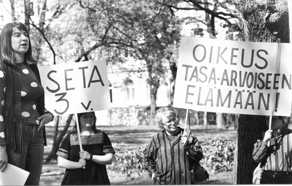 Ihmisiä mielenosoituksessa - vasemmalla Marita Kuokkanen puhumassa, keskellä kolme henkilöä, jotka pitävät kylttejä, joissa lukee "Seta 3v." ja "Oikeus tasa-arvoiseen elämään!"