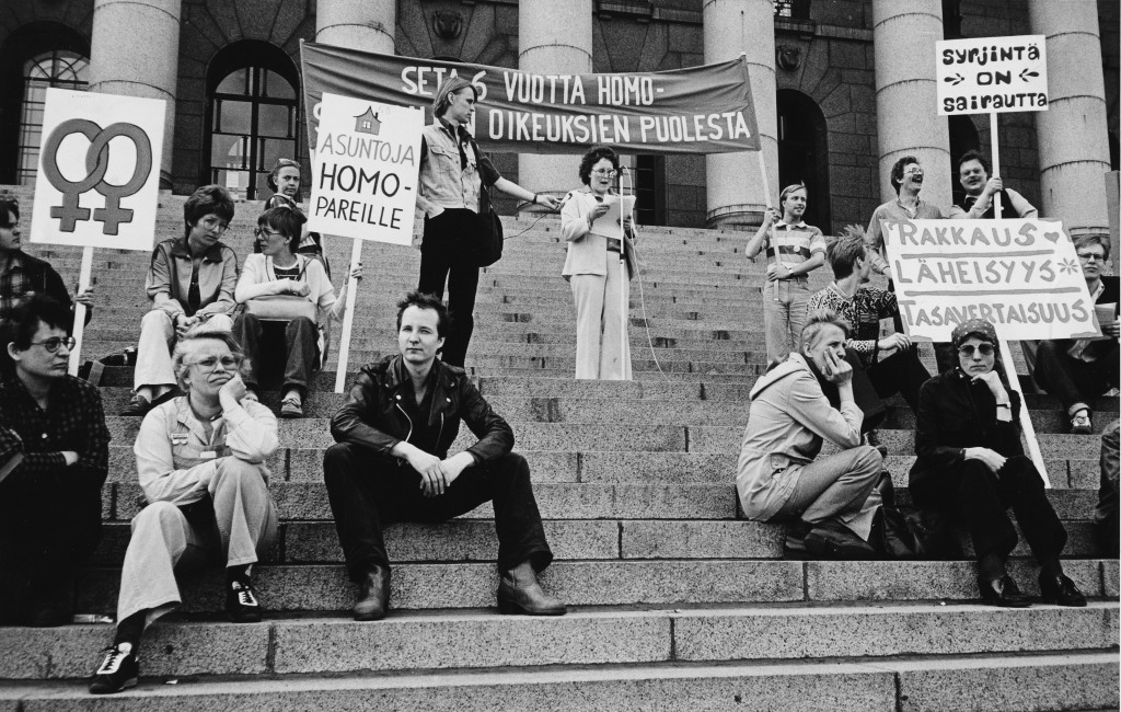 Demonstranter på riksdagshusets trappor. Skyltarna pryds av dubbla venustecken samt texterna "Lägenheter för homosexuella par", "Diskriminering är sjukdom" och "Kärlek, intimitet, jämlikhet." På en stor banderoll i mitten står “Seta sex år för homosexuella rättigheter”, en del av banderolltexten skyms av de människor som står framför den.