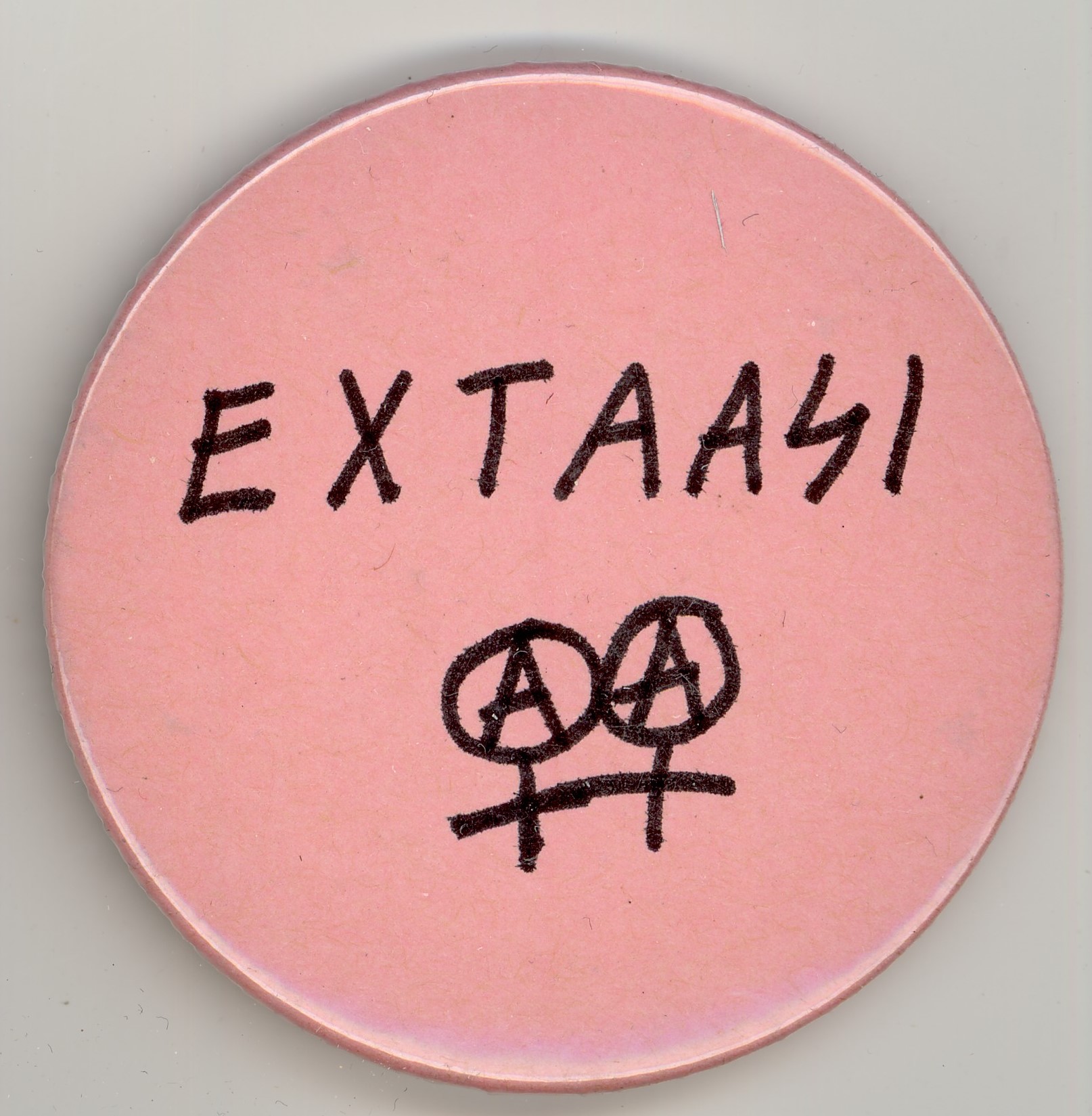 Vaaleanpunainen pinssi, jossa lukee mustalla käsinkirjoitetulla tekstillä Extaasi. Tekstin alla tuplanarttumerkit, joiden sisällä A-kirjain.