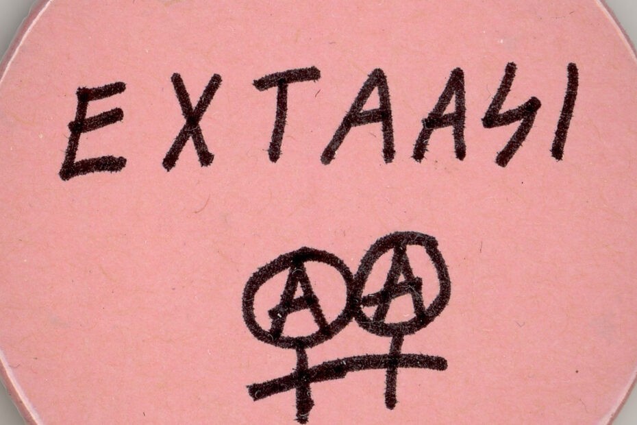 Vaaleanpunainen pinssi, jossa lukee käsin kirjoitettu teksti Extaasi ja alla kaksoisnarttumerkki, jossa ympyröiden sisällä A-kirjain.