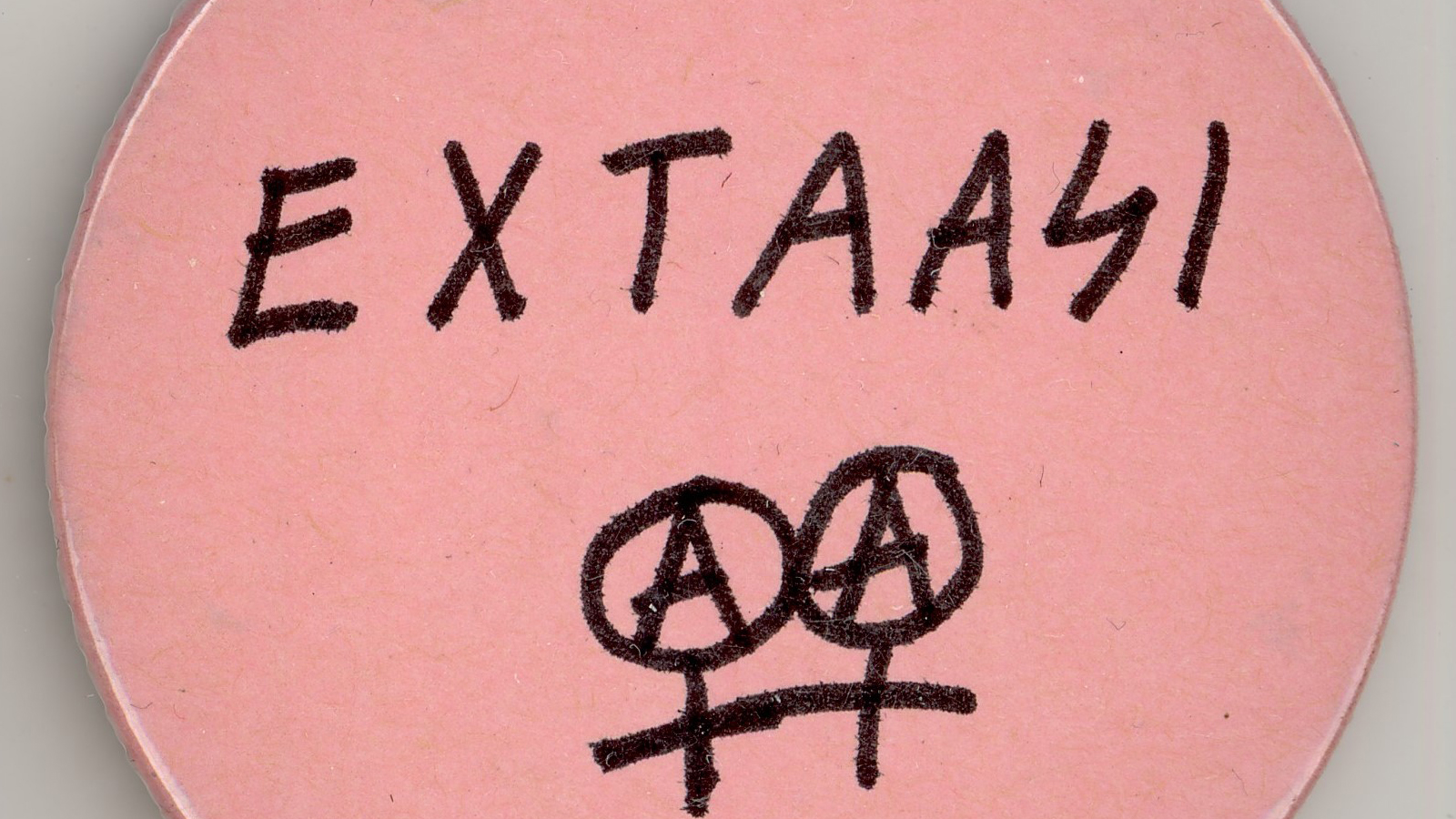 Vaaleanpunainen pinssi, jossa lukee käsin kirjoitettu teksti Extaasi ja alla kaksoisnarttumerkki, jossa ympyröiden sisällä A-kirjain.