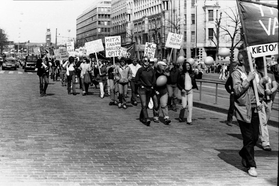 Mustavalkokuva mielenosoituksesta Mannerheimintiellä Ylioppilastalon edeustalla. Marssijat pitelevät kylttejä ja marssin takana näkyy autoja pysähtyneenä