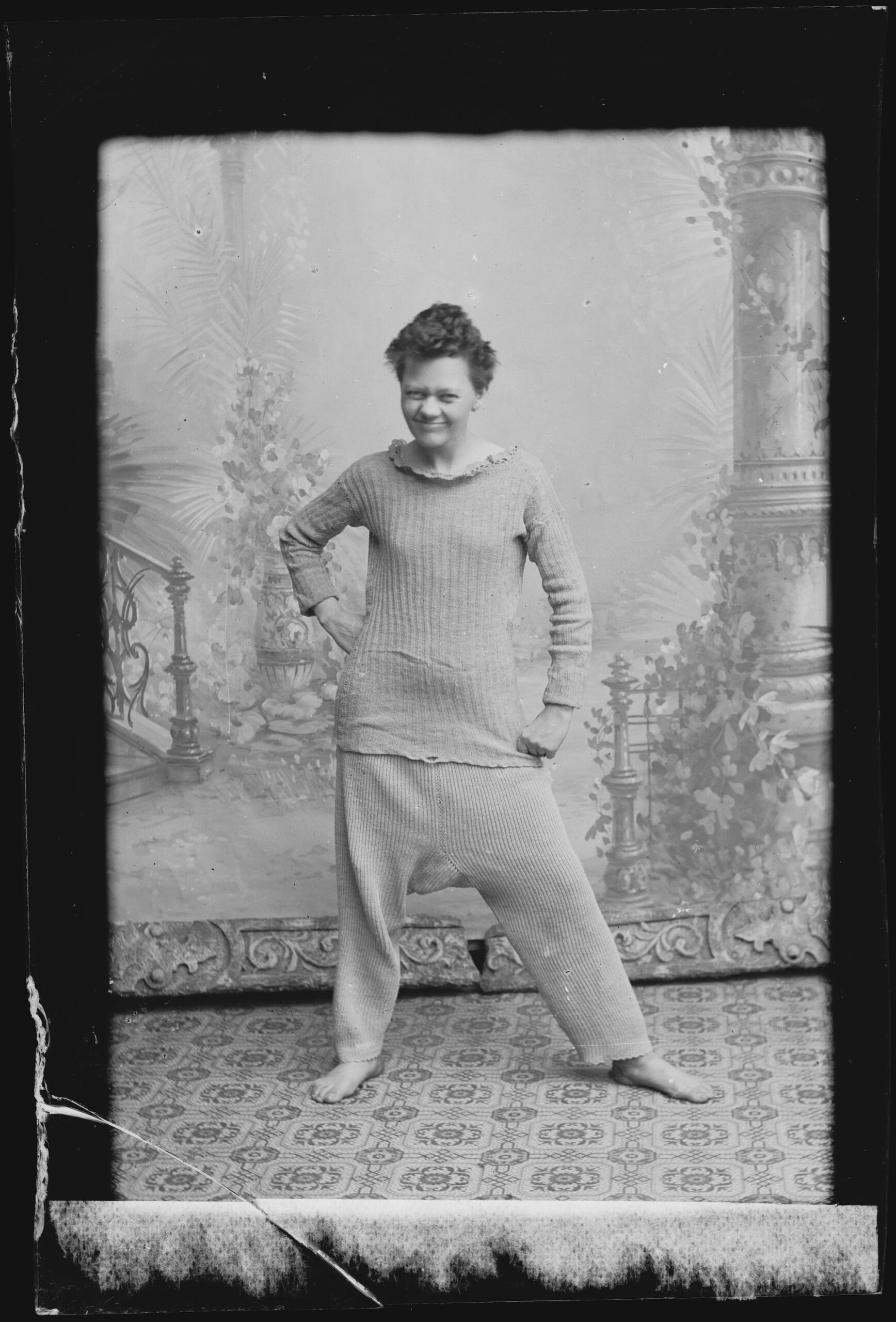 Marie Høeg mustavalkokuvassa vanhassa valokuvastudiossa maisemakankaan edessä. Hänellä on yllään pyjamalta näyttävä vaate ja hänen ilmeensä on vitsikäs.
