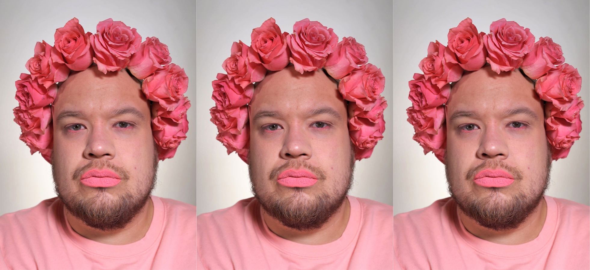 Valkoihoinen henkilö, jolla on vaaleanpunaisista ruusuista koostuva seppele, vaaleanpunaista huulipunaa, parta, sekä vaaleanpunainen paita.
