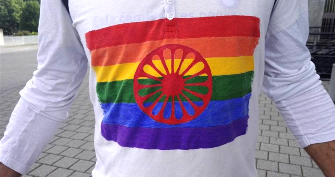 Henkilö valkoisessa t-paidassa, jossa romanilipun punainen kärrynpyöräsymboli sateenkaaripohjalla.