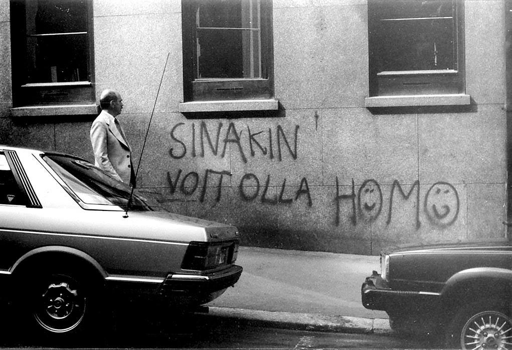 Mustavalkokuva rakennuksen kivijalasta, johon on kirjoitettu graffitilla "sinäkin voit olla homo". Homo-sanan o-kirjaimissa on hymynaamat. Ohikulkija katsoo graffitia.