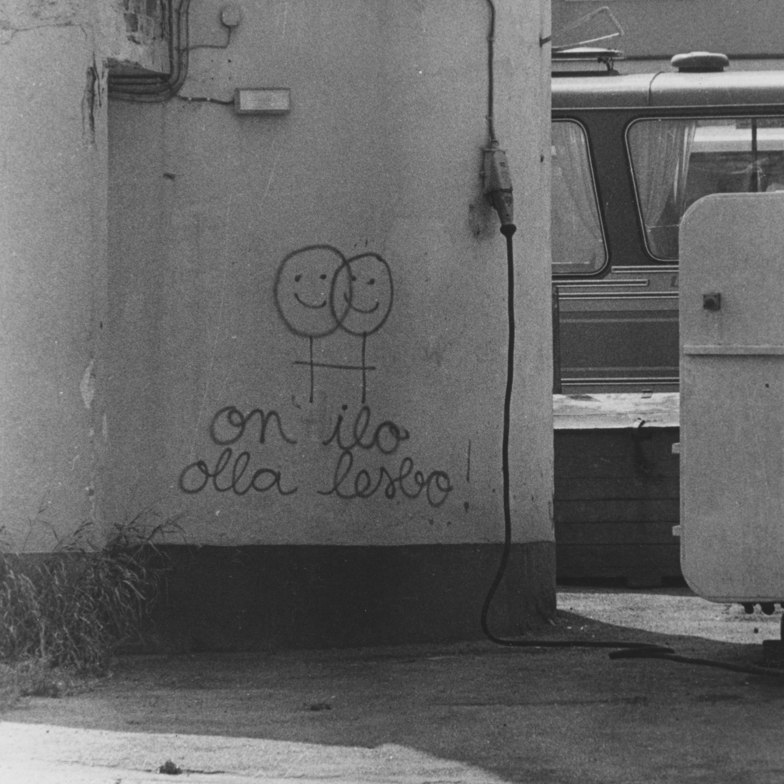 Mustavalkokuva seinään tehdystä graffitista, jossa tuplanarttumerkki hymynaamoilla ja alla teksti "on ilo olla lesbo!"