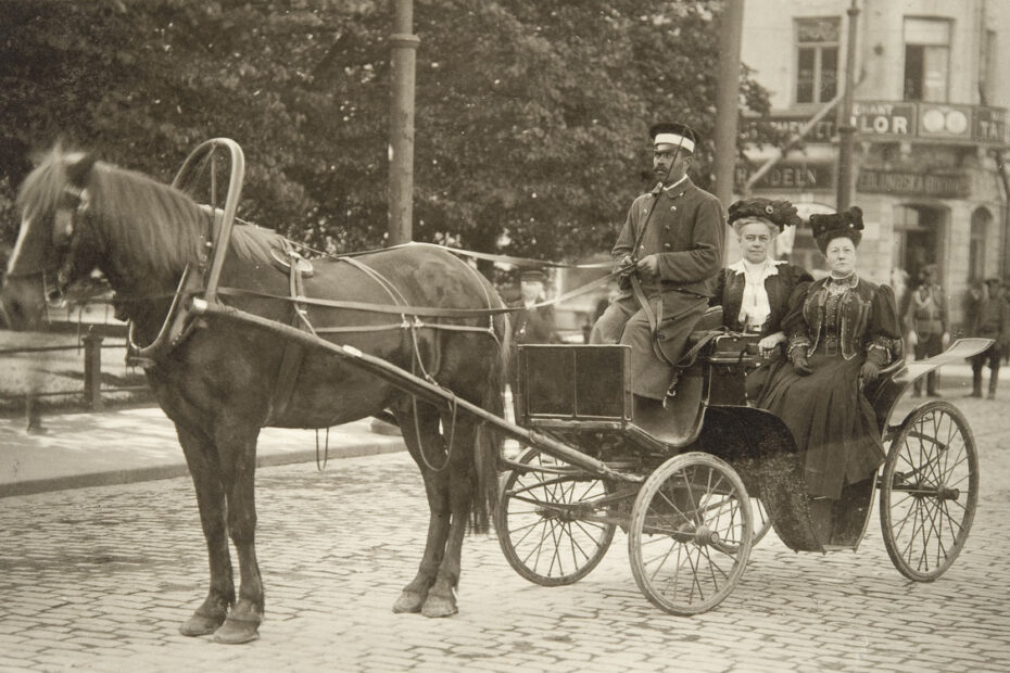Mustavalkokuva kahdesta naisesta avoimessa hevosvaunussa mukulakivetyllä kadulla.