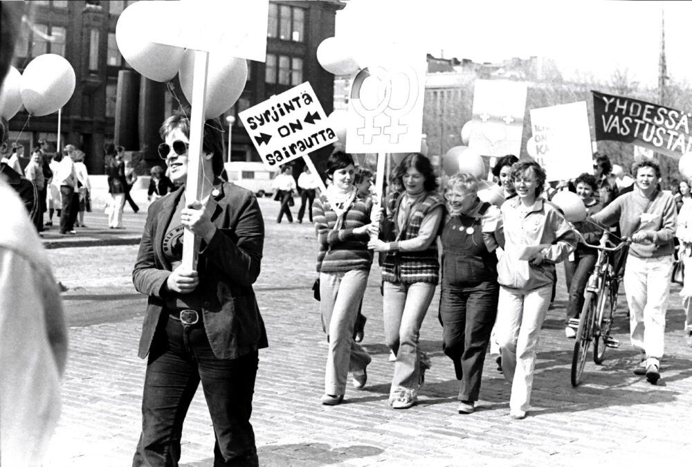 Mieltä osoittavia iloisia naisia. Kyltissä teksti "syrjintä on sairautta".