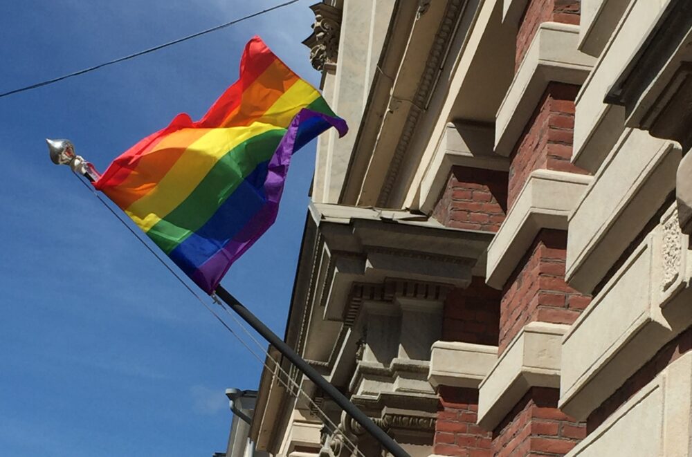 Sateenkaarilippu liehuu, taustalla näkyy sininen taivas ja SKS:n rakennuksen seinä.