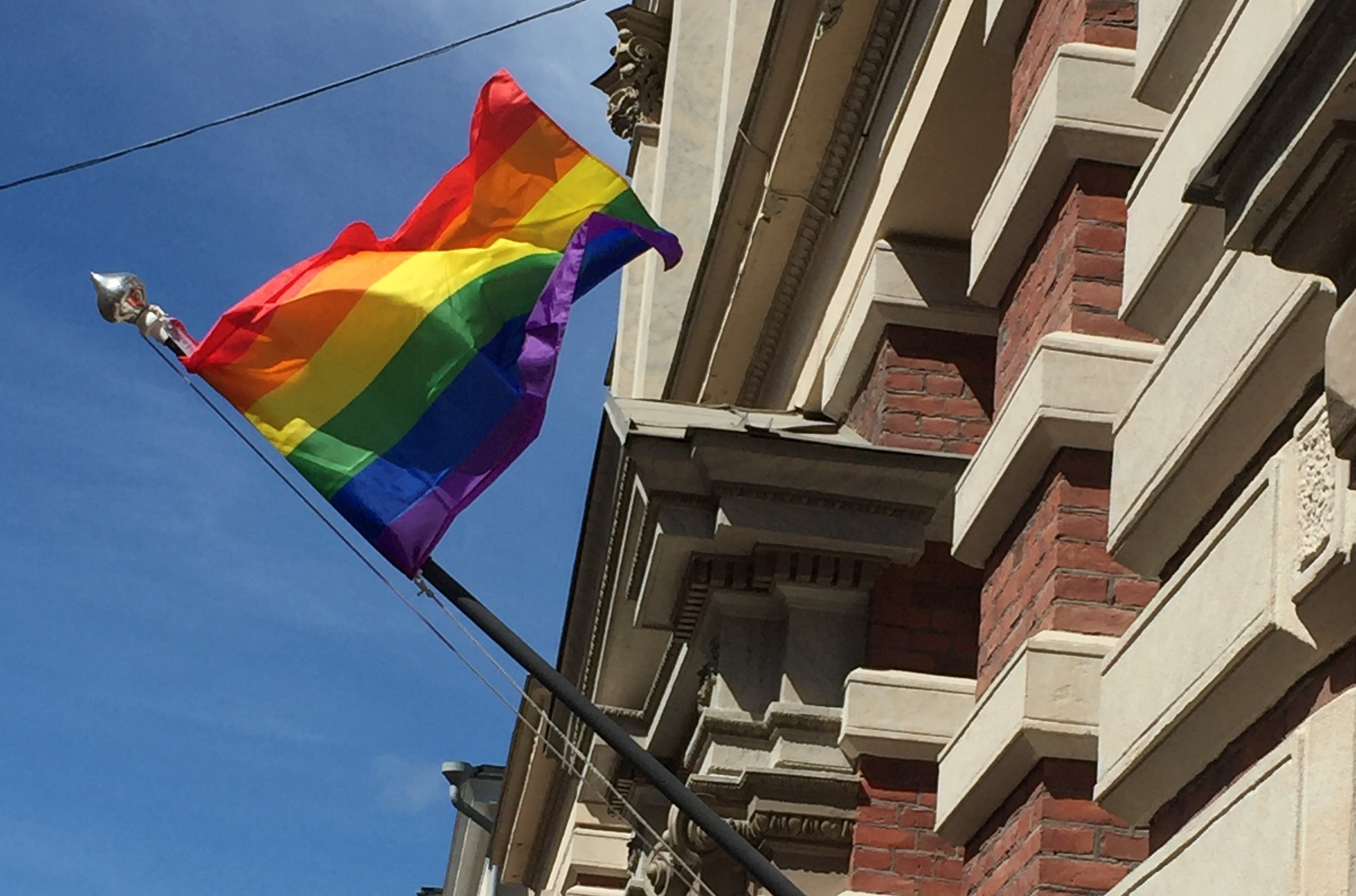 Sateenkaarilippu liehuu, taustalla näkyy sininen taivas ja SKS:n rakennuksen seinä.