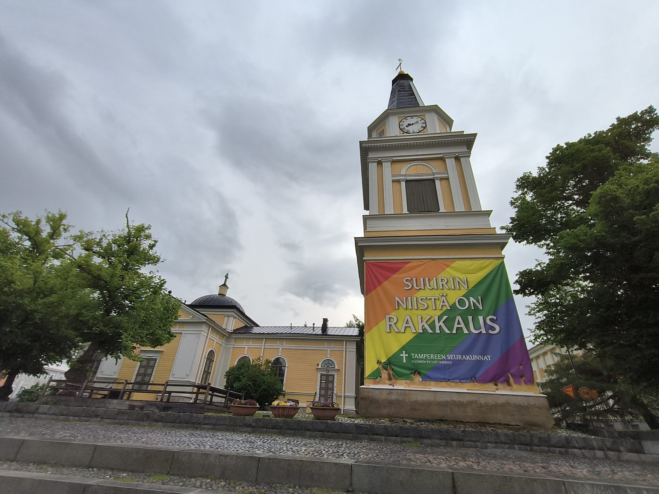 Pilvisenä kesäpäivänä Tampereen Vanhan kirkon kirkkotapuli, johon on ripustettuna sateenkaaribanderolli, jota on poltettu alareunasta. Banderollissa lukee "Suurin niistä on rakkaus."