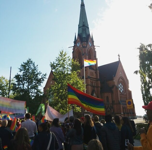 Aurinkoinen kuva Uumajan kirkosta ja Pride-marssijoista sateenkaarilippujen kera