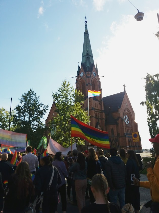 Aurinkoinen kuva Uumajan kirkosta ja Pride-marssijoista sateenkaarilippujen kera